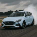 A Hyundai i30 N hot hatch in Performance Blue drifts around a racetrack corner, emitting white tire smoke, with a blurred track and overcast sky in the background.