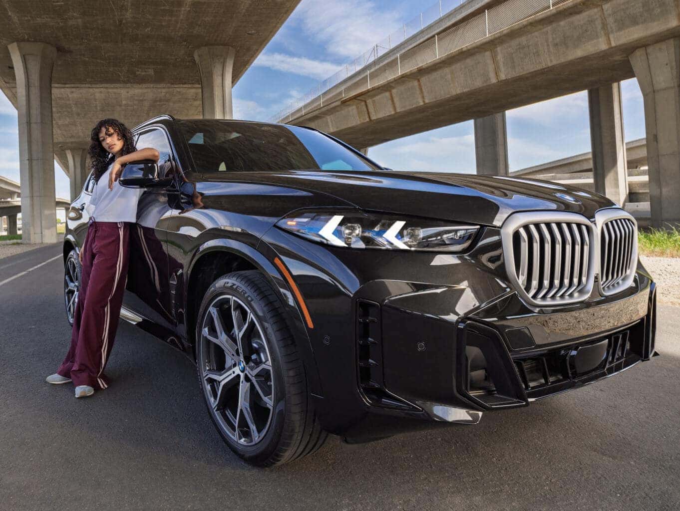 Wide angle view of BMW X5 parked under a freeway with woman leaning on passenger door
