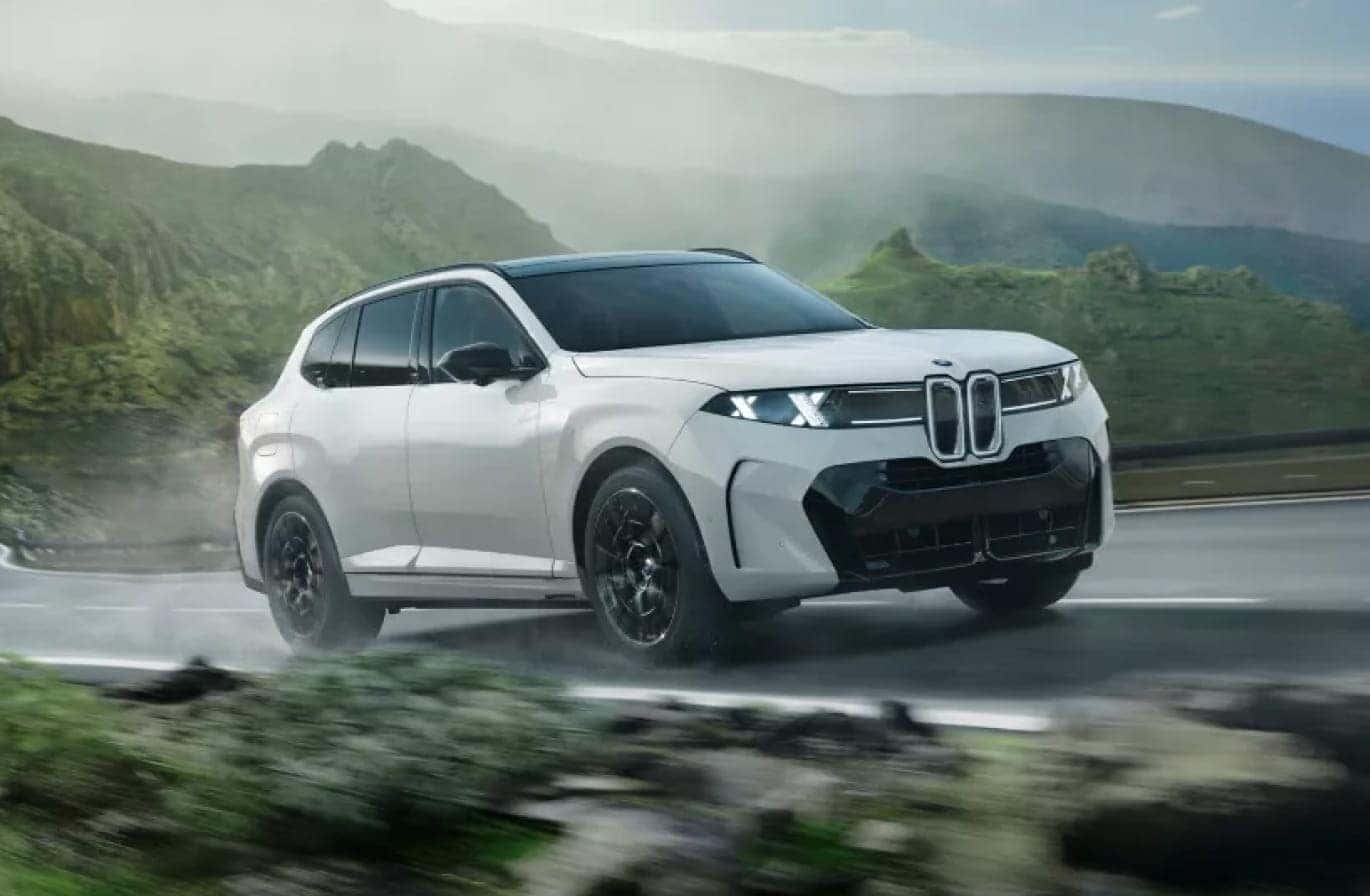 White BMW X5 driving on a wet mountain road with lush green hills and misty scenery in the background.