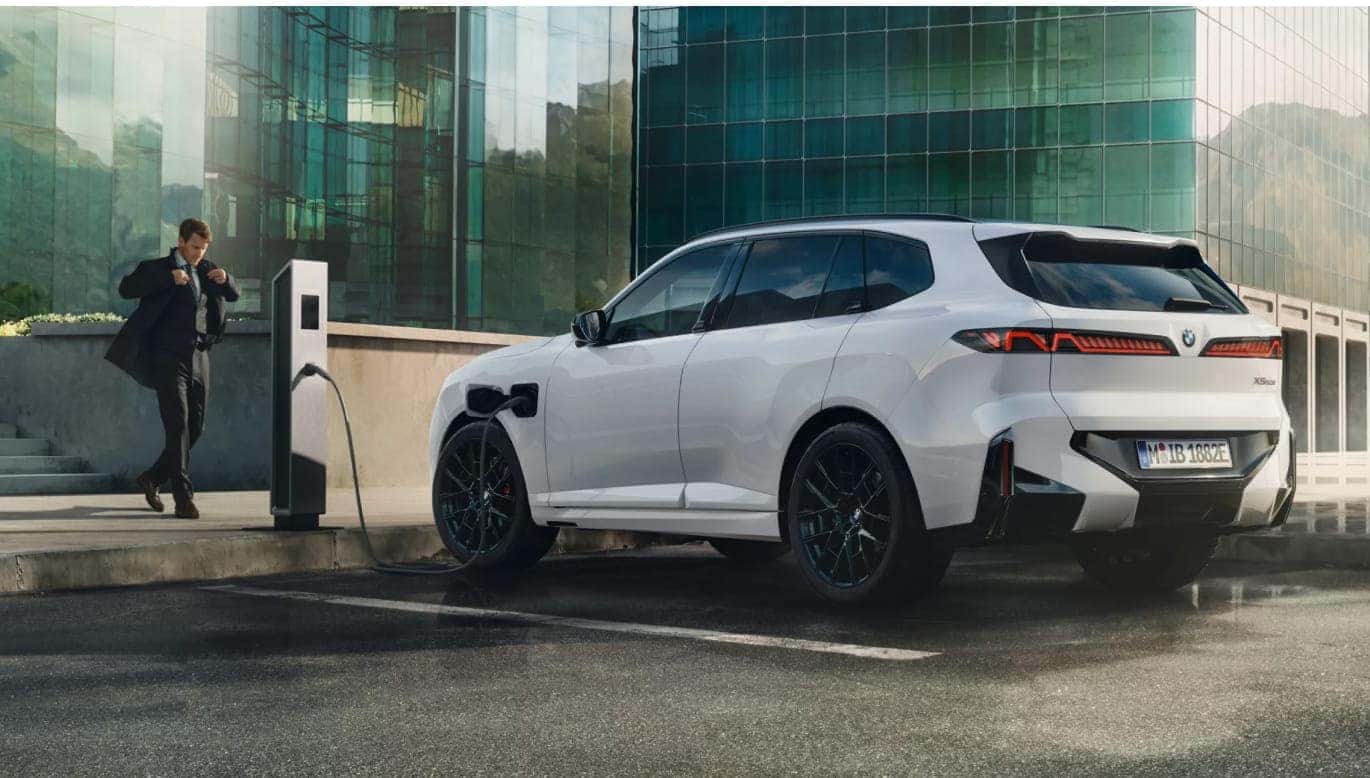 White BMW X5 plugged into a charging station outside a modern glass building with a person walking nearby.