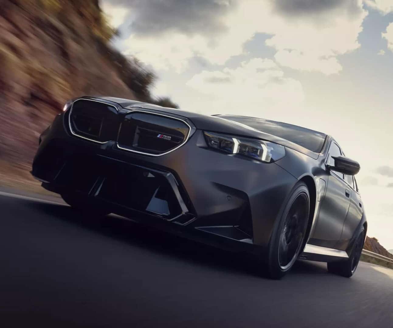 Low angle front action shot of BMW 5 Series sedan speeding down a sunny remote highway.