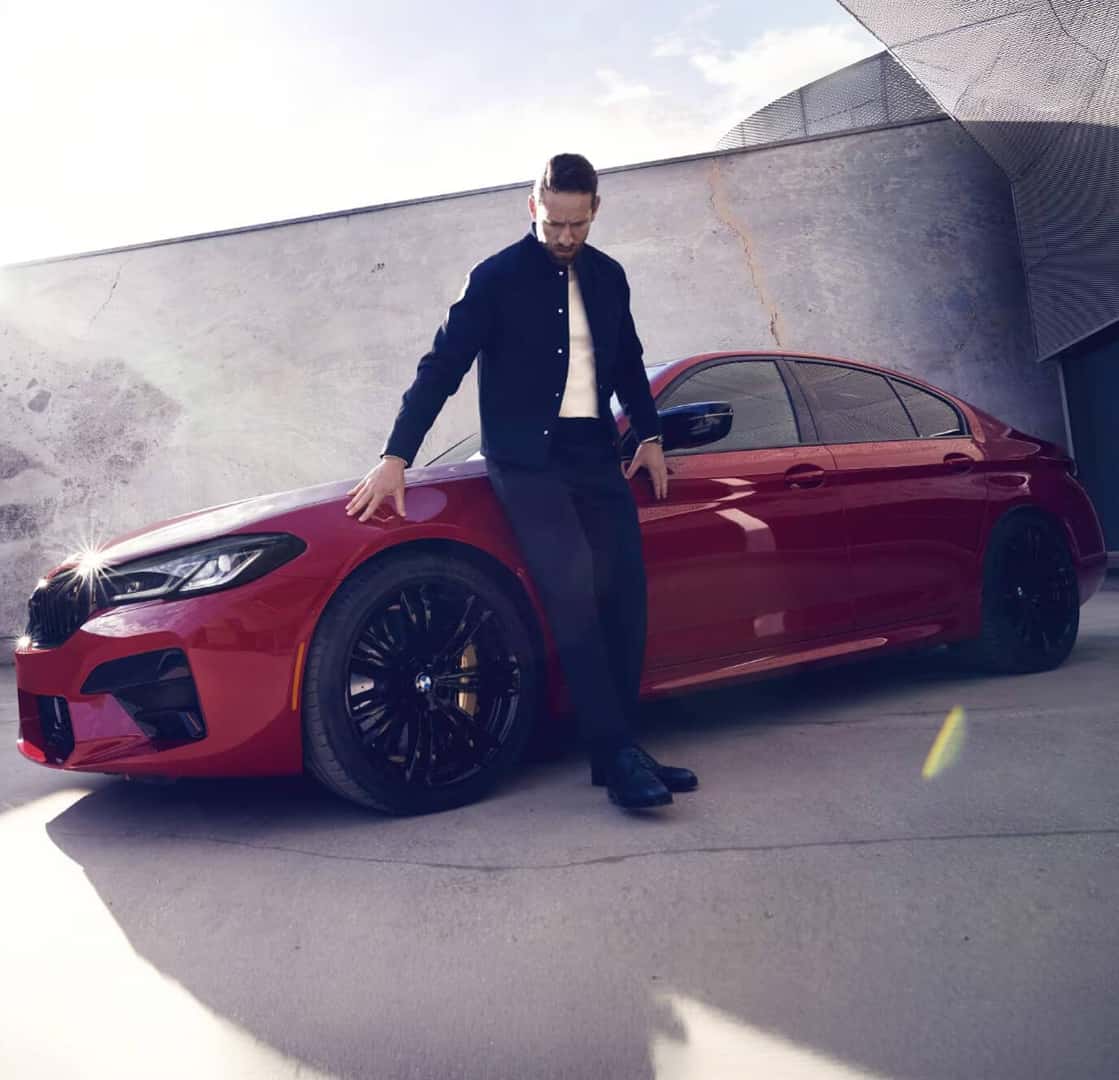 Side profile view of a man leaning against a 2026 BMW M5 in Toronto Red Metallic outside a modern concrete structure.