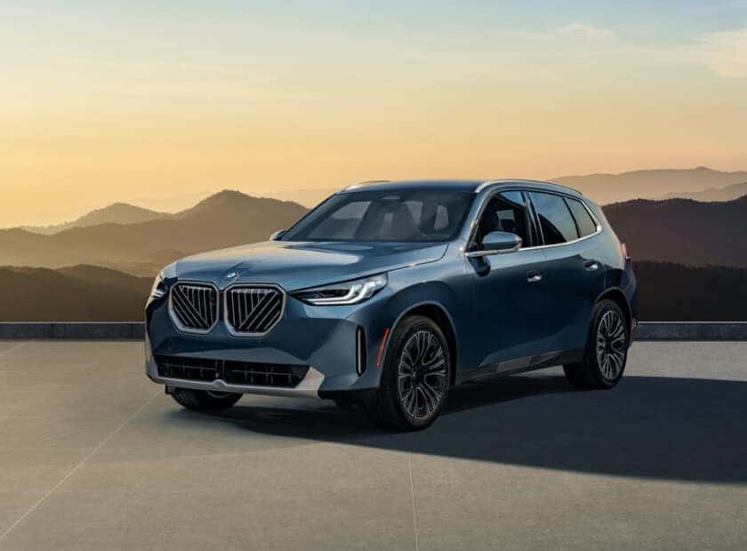 Front three-quarter view of blue BMW X3 parked at sunset with mountains in background.