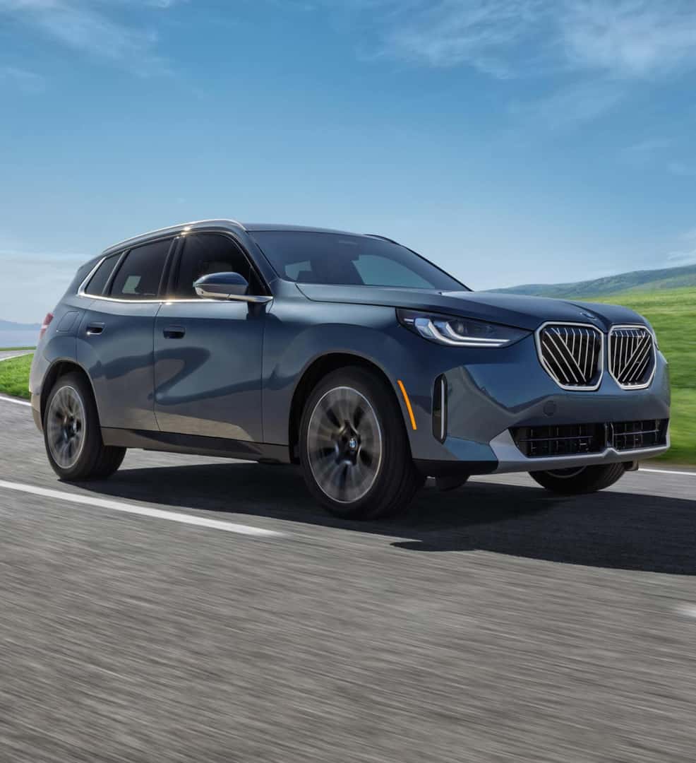Front three-quarter view of blue BMW X3 driving on a road near green hills and ocean.