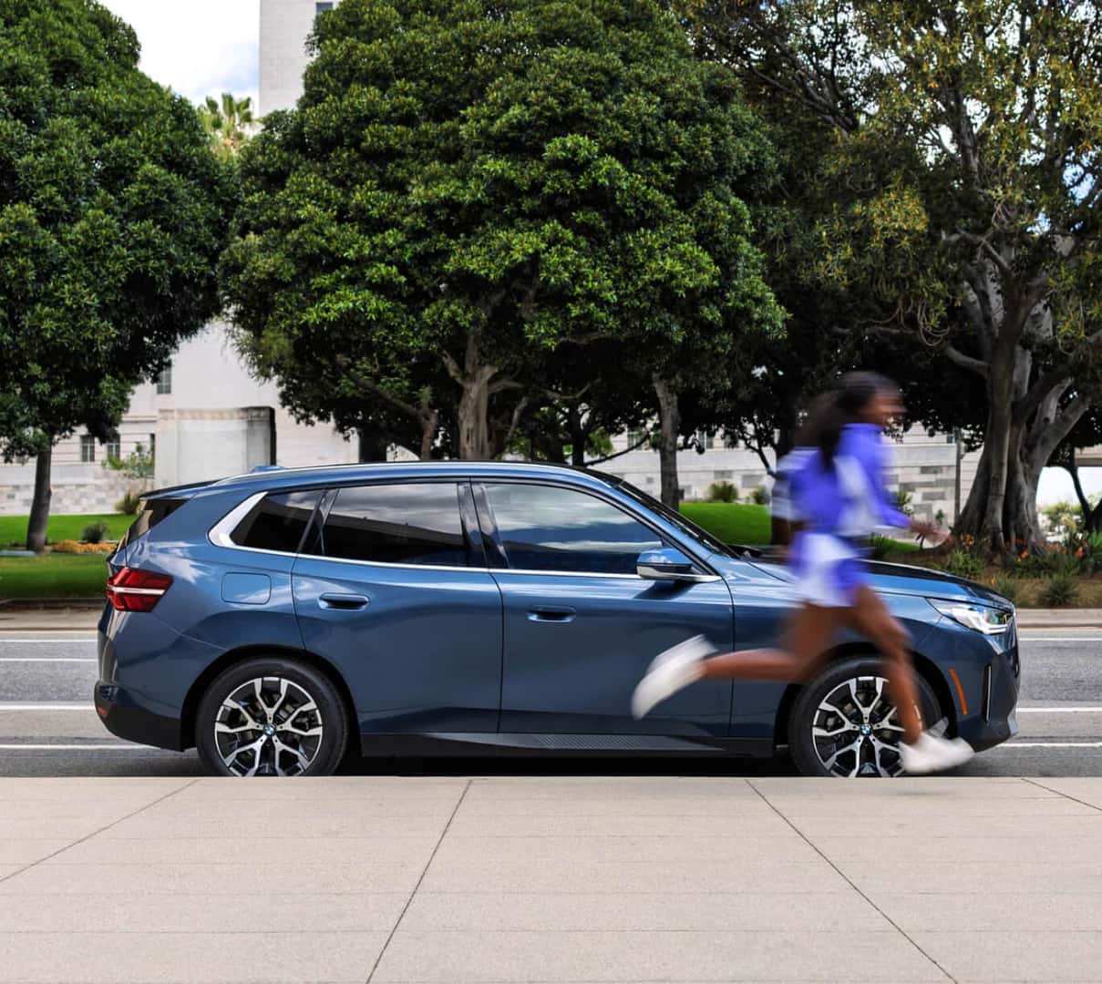 Side profile view of blue BMW X3 parked on city sidewalk with trees background.
