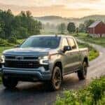 A powerful 2026 Chevrolet Silverado 1500 in metallic slate grey driving along a winding rural road in South-Central Pennsylvania, low-angle three-quarter front perspective to emphasize a dominant stance, rolling green hills and traditional red barns with stone foundations in the soft-focus background, early morning golden hour lighting with a light valley mist, vibrant emerald pastures and historic limestone fences, hyper-realistic automotive photography style, crisp details on the textured grille and signature LED daytime running lights, heavy-duty utility aesthetic, reflections of the Appalachian landscape on the polished bodywork, warm cinematic atmosphere, lush vegetation, professional color grading, 8k resolution.