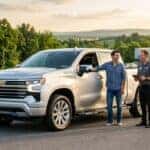 A man in casual attire talking to a dealership representative next to a silver Chevrolet Silverado pickup truck.