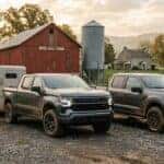 Cinematic wide shot of a 2026 Chevrolet Silverado 1500 and a Ford F-150 parked side-by-side on a traditional South-Central Pennsylvania farm, low-angle perspective emphasizing ruggedness, the Silverado is the focal point hitched to a heavy livestock trailer on a gravel staging area, setting includes a large weathered red wooden barn, a tall silver grain silo, and a historic limestone farmhouse in the distance, rolling green pastures and misty Appalachian hills under a soft morning haze, golden hour sunlight filtering through oak trees, realistic textures of high-strength steel truck beds and muddy all-terrain tires, earthy color palette of deep greens, rustic red, and metallic charcoal, high-detail automotive photography, serene and industrious agricultural mood.