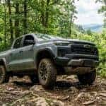 A 2026 Chevrolet Colorado Trail Boss, mid-size off-road pickup truck, rugged slate-gray finish, high-clearance stance, threading through a narrow and technical logging road in Michaux State Forest, South Mountain terrain, south-central Pennsylvania, sharp rocky ridges and dense timber forest in the background, realistic June afternoon, bright dappled sunlight filtering through a thick canopy of oak and maple trees, lush green ferns and mountain laurel lining the trail, subtle mud splatters on the widened wheel arches, low-angle shot focusing on the underbody transfer case shield and all-terrain tires, realistic outdoor scouting mood, crisp and detailed forest textures, hazy Appalachian blue ridges in the far distance, natural and earthy color palette, cinematic realism, 8k resolution.