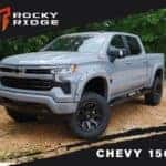A gray Chevy 1500 Rocky Ridge lifted pickup truck on a dirt path with custom wheels.