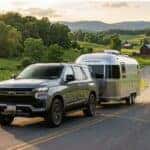Grey Chevrolet SUV towing a silver Airstream trailer along a winding country road past green hills and a red barn.