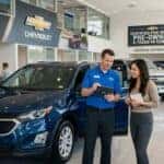 A Chevrolet salesman showing a blue SUV to a female customer inside a brightly lit car dealership showroom. (Representative image for illustration purposes only. Actual vehicle may vary based on trim and configuration.)