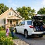 A woman packs backpacks into the open trunk of a white SUV in a driveway while two children stand nearby. (Representative image for illustration purposes only. Actual vehicle may vary based on trim and configuration.)