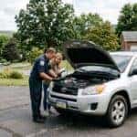 A mechanic inspects the engine of a silver SUV in a driveway while a woman holding a clipboard watches. (Representative image for illustration purposes only. Actual vehicle may vary based on trim and configuration.)