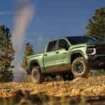 Green Chevrolet Silverado pickup truck driving over rocky off-road terrain against a blue sky with pine trees.