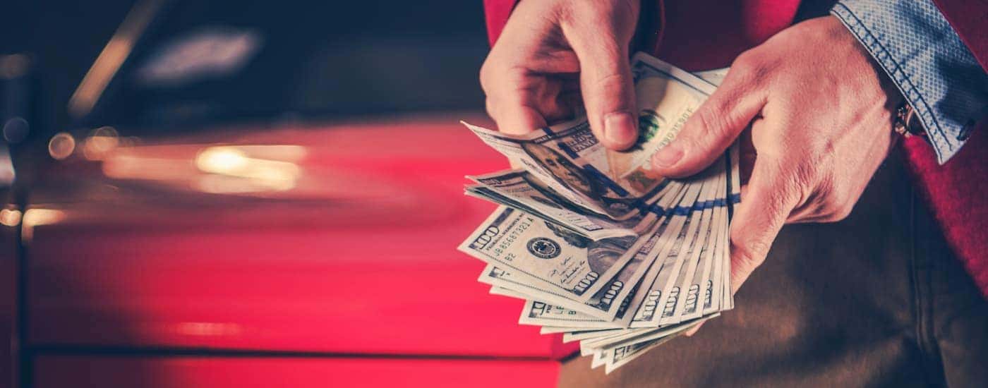 A person is shown holding a hand full of cash in front of a car.