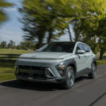 Light green 2025 Hyundai Kona driving past a tree-lined road with motion blur