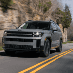 Gray 2025 Hyundai Santa Fe Hybrid driving on a scenic mountain road near cliffside trees
