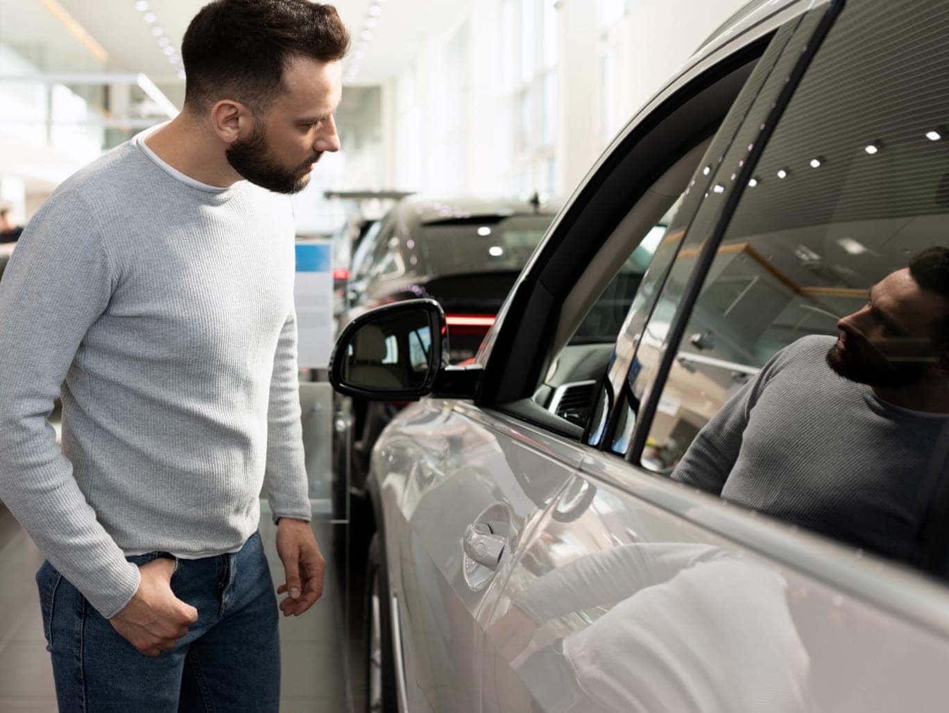 Text: Man inspecting the side of a white SUV inside a bright dealership showroom with other vehicles in the background.