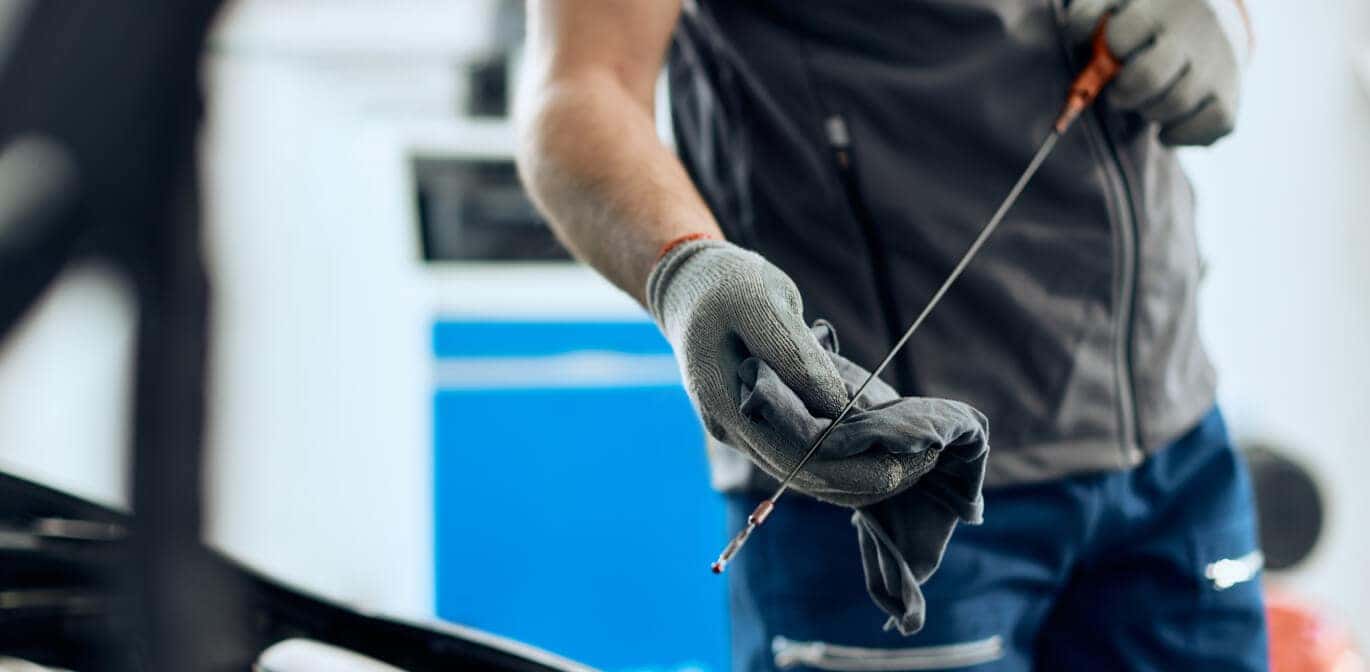 Technician checking a car engine oil dipstick under an open hood in a service bay.
