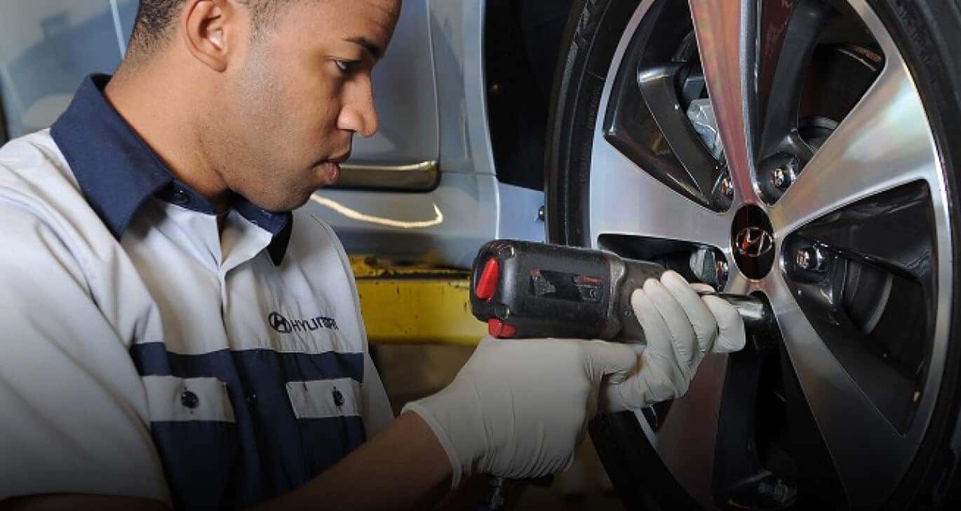 Hyundai technician using an impact wrench on a light blue vehicle wheel in a service garage.