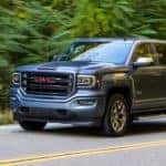 A grey 2016 GMC Sierra, popular among used cars near me, is driving on a tree-lined road.