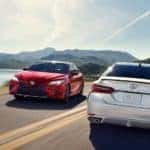 A white 2019 Toyota Camry, which is popular among used cars, is driving past a red one on a road next to a lake.