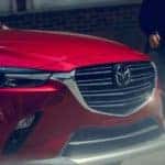 The front of a red 2019 Mazda CX-3 is shown on a city street with a woman walking toward it.
