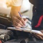 A mechanic is filling out paperwork on a clipboard over an engine bay.