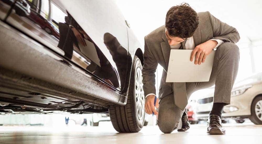 A man with a clipboard is kneeling to look under a black car.