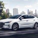 A white 2020 Hyundai Elantra is shown driving on an open road past a city.