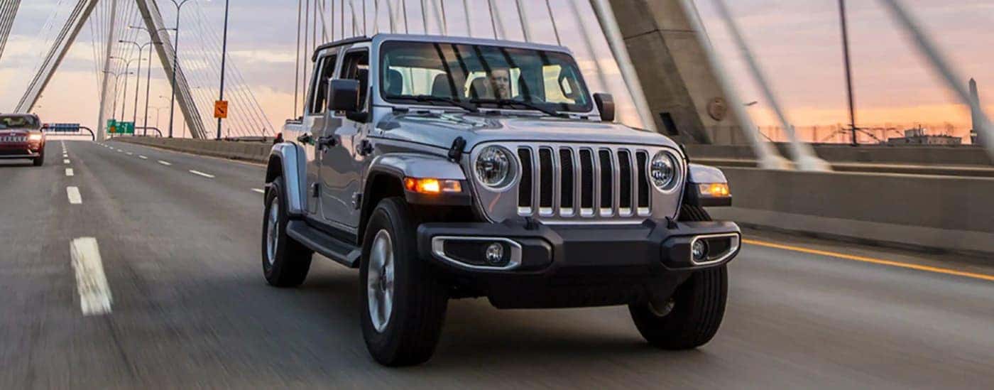 A silver 2021 Jeep Wrangler Unlimited is shown driving over a bridge at sunset.