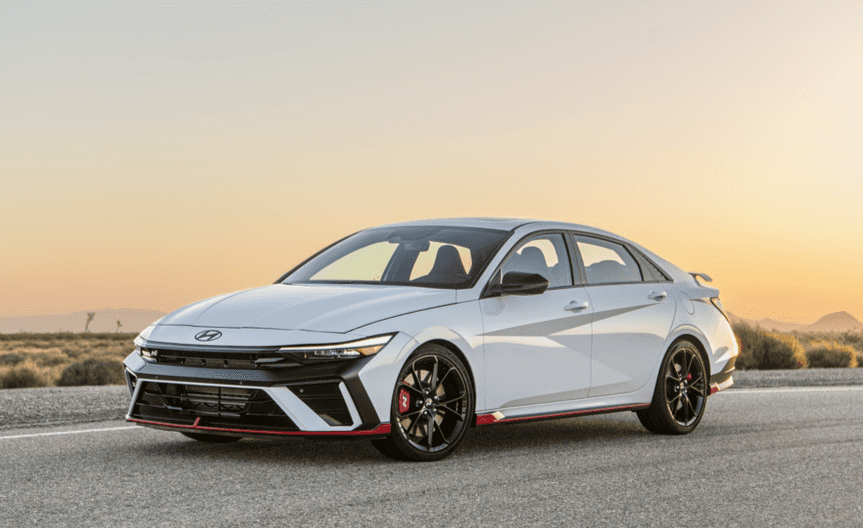 A 2025 Hyundai Elantra N drives along a scenic road during a vibrant sunset, showcasing its sleek design and performance.