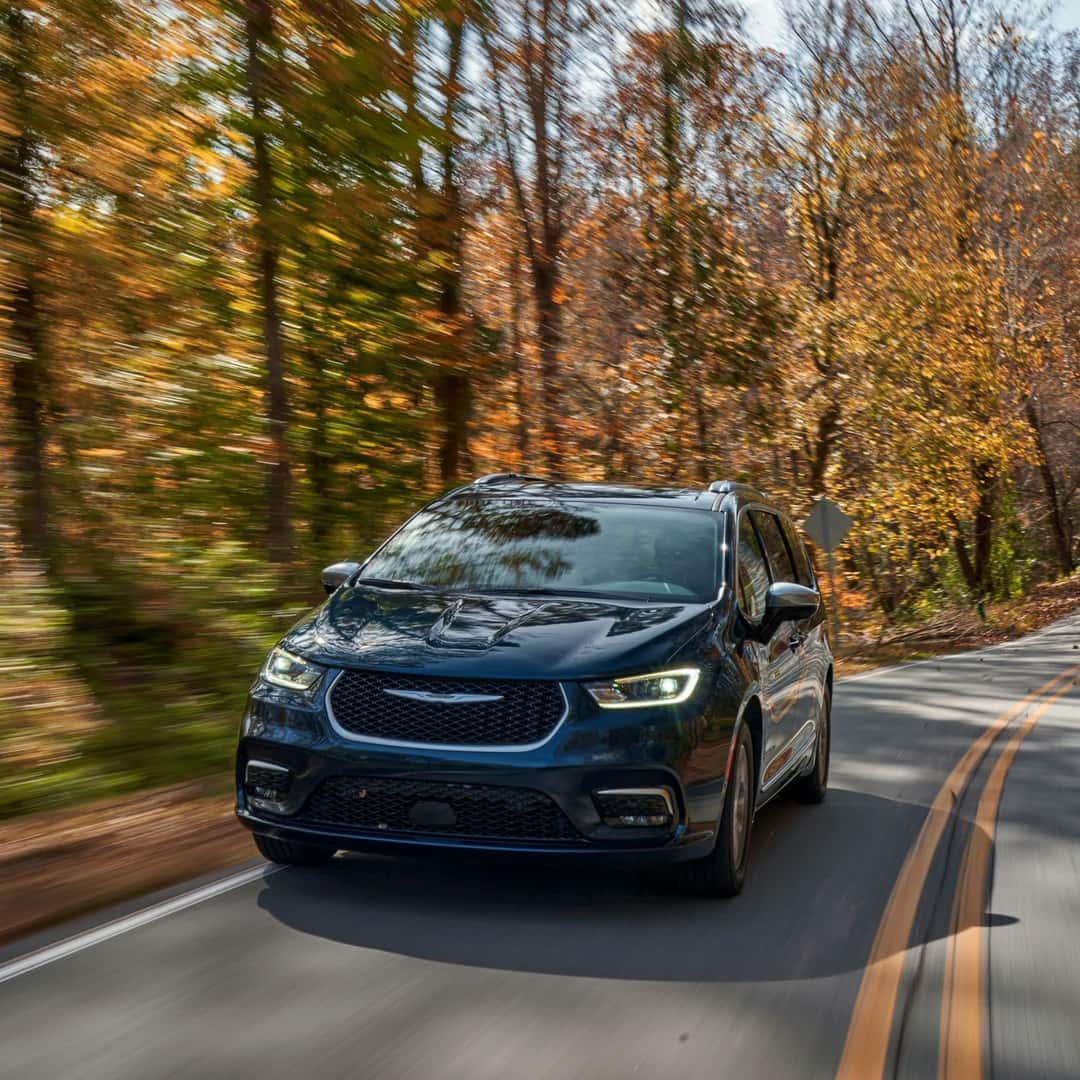 2025 Pacifica Hybrid (RU) Pinnacle in Fathom Blue Pearl with lit headlamps driving on a hilly, tree-lined road in the Fall.
