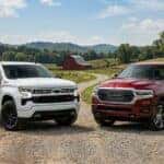 A white 2025 Chevy Silverado 1500 and a red 2025 Ram 1500 pickup truck parked together on a country road with an Indiana farm and red barn in the background.
