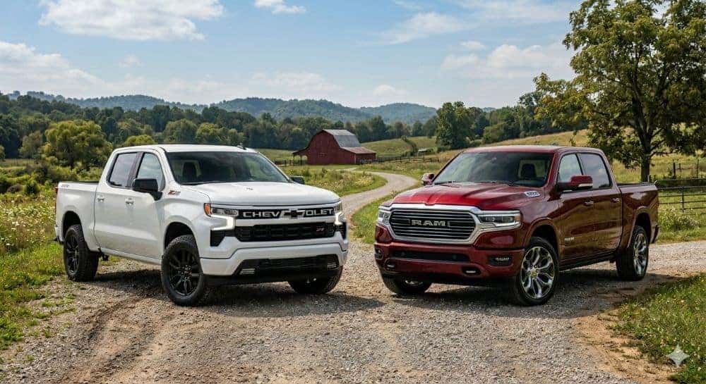 A white 2025 Chevy Silverado 1500 and a red 2025 Ram 1500 pickup truck parked together on a country road with an Indiana farm and red barn in the background.