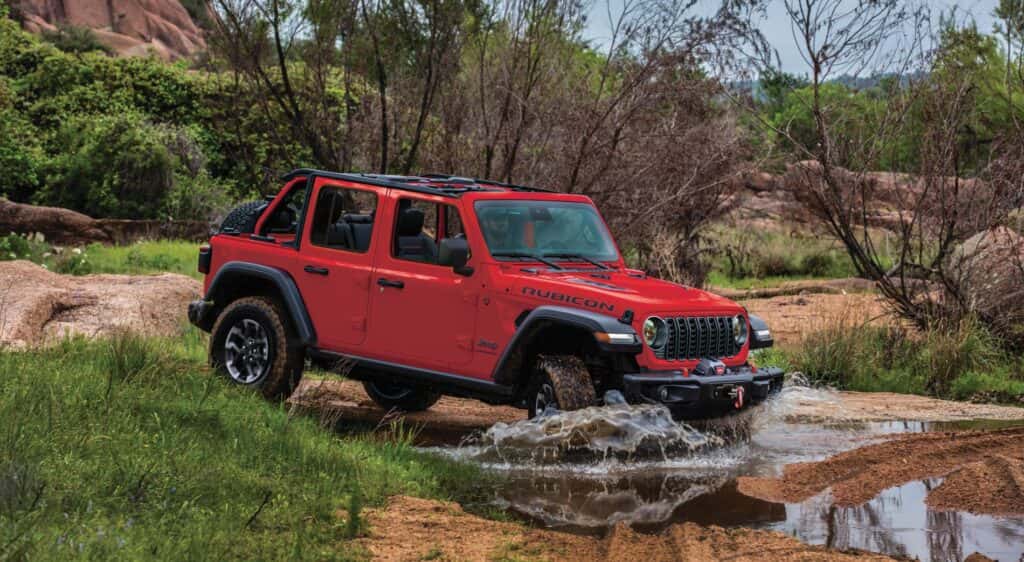 2026 Wrangler Rubicon in Firecracker Red driving into a shallow creek, with tree