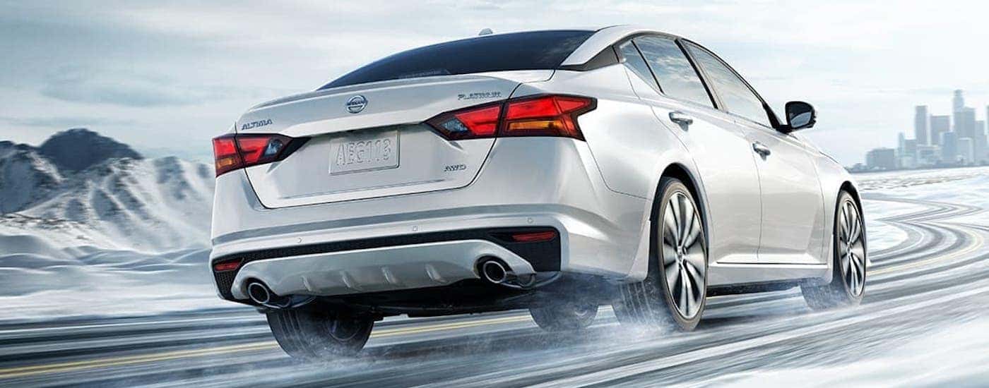 A white 2021 Nissan Altima is shown from behind while driving on a snowy road towards a city.