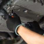 Gloved hands are removing the oil plug under a car.