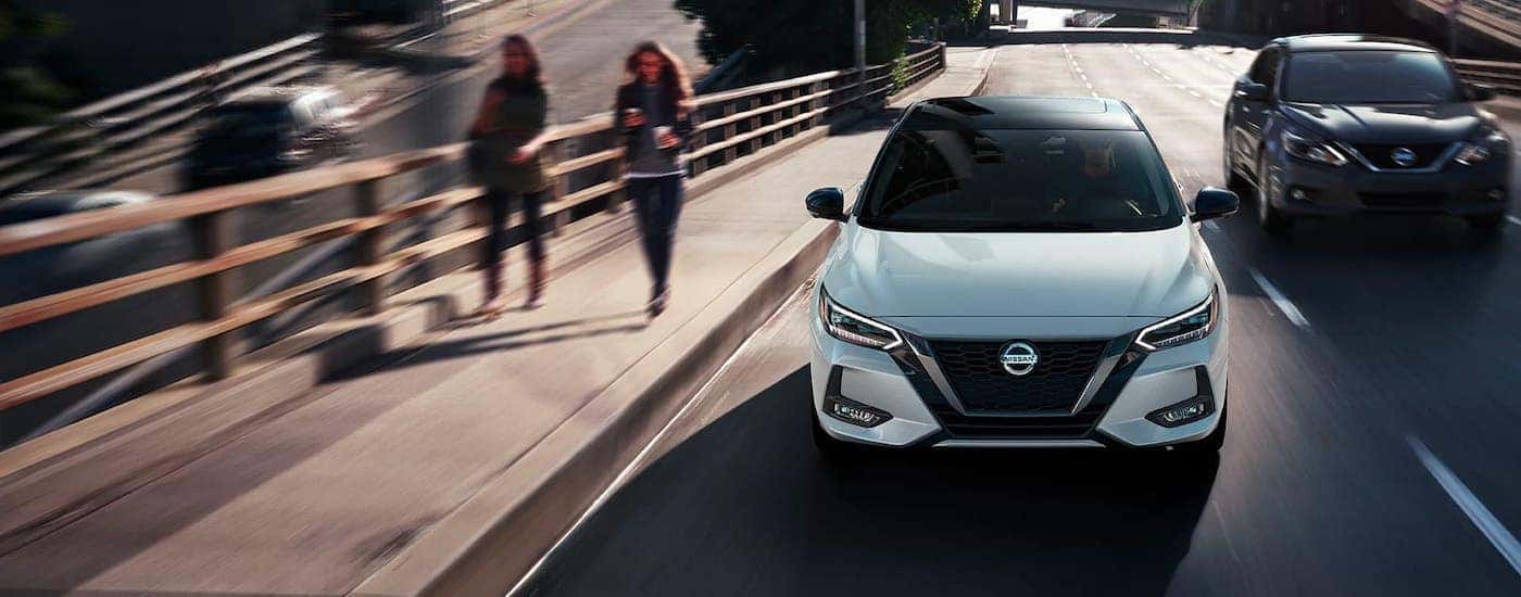 One of the most popular new cars in stock, a white 2021 Nissan Sentra, is shown driving down the street next to pedestrians.