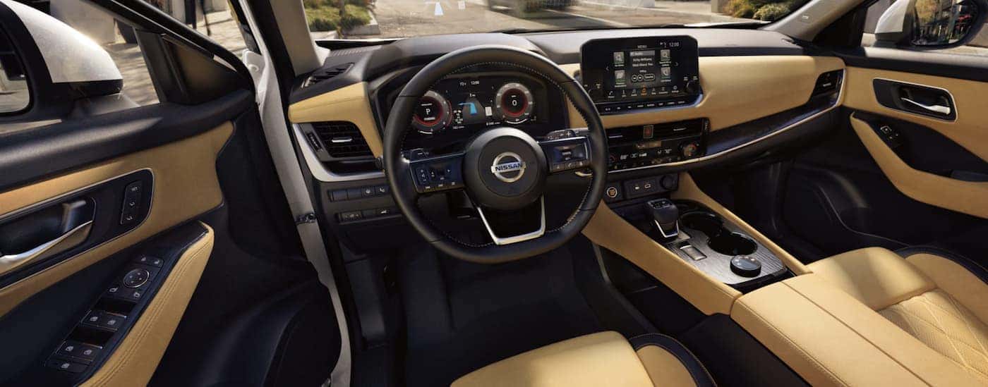 The black and tan interior of a 2021 Nissan Rogue shows the steering wheel and infotainment screen.