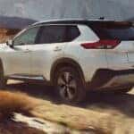 A white 2023 Nissan Rogue is shown off-roading on a dirt path in the mountains.