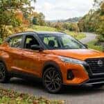 A high-quality editorial photograph of a 2025 Nissan Kicks parked along a scenic paved suburban road in Middle Tennessee during early autumn, framed by gently rolling green hills and turning oak trees, three-quarter front view capturing the modern compact crossover design, crisp natural daylight casting soft reflections on the metallic painted body, driver door slightly open revealing a sleek dark interior with a modern digital dashboard, quiet rural suburban environment, realistic colors, rich detail. No text, no logos other than natural vehicle badging, no identifiable faces. (Representative image for illustration purposes only. Actual vehicle may vary based on trim and configuration.)