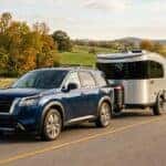 A professional lifestyle photograph of a 2025 Nissan Pathfinder in a sleek deep blue finish, smoothly towing a modern lightweight camper trailer along a scenic two-lane country road in Middle Tennessee. Captured from a dynamic low three-quarter angle, emphasizing the vehicle sturdy stance and towing capability. The surrounding landscape features gently rolling green hills, wooden farm fences, and scattered hardwood trees displaying vibrant early autumn foliage in warm amber and gold tones. Soft late-afternoon sun casts long gentle shadows and highlights on the metallic car body, creating an authentic, inspiring family travel scene. No text, no logos other than natural vehicle badging, no identifiable faces. (Representative image for illustration purposes only. Actual vehicle may vary based on trim and configuration.)