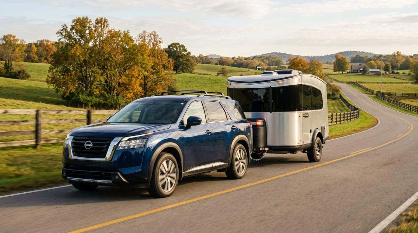 A professional lifestyle photograph of a 2025 Nissan Pathfinder in a sleek deep blue finish, smoothly towing a modern lightweight camper trailer along a scenic two-lane country road in Middle Tennessee. Captured from a dynamic low three-quarter angle, emphasizing the vehicle sturdy stance and towing capability. The surrounding landscape features gently rolling green hills, wooden farm fences, and scattered hardwood trees displaying vibrant early autumn foliage in warm amber and gold tones. Soft late-afternoon sun casts long gentle shadows and highlights on the metallic car body, creating an authentic, inspiring family travel scene. No text, no logos other than natural vehicle badging, no identifiable faces. (Representative image for illustration purposes only. Actual vehicle may vary based on trim and configuration.)