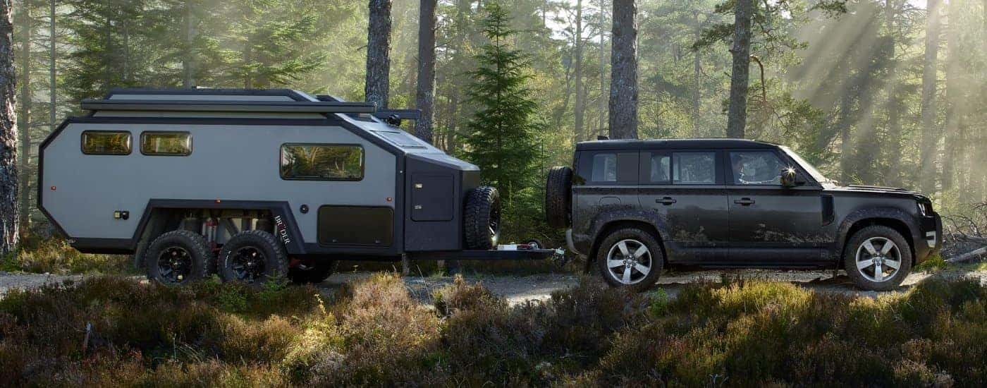 A black 2025 Land Rover Defender 110 is towing a camper in a forest.