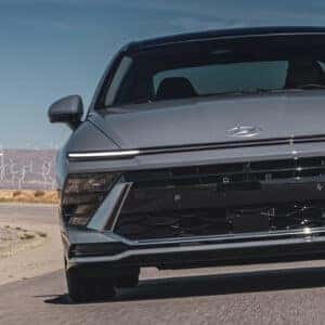 Front view of the 2024 Hyundai Sonata driving on an open road with wind turbines in the background.