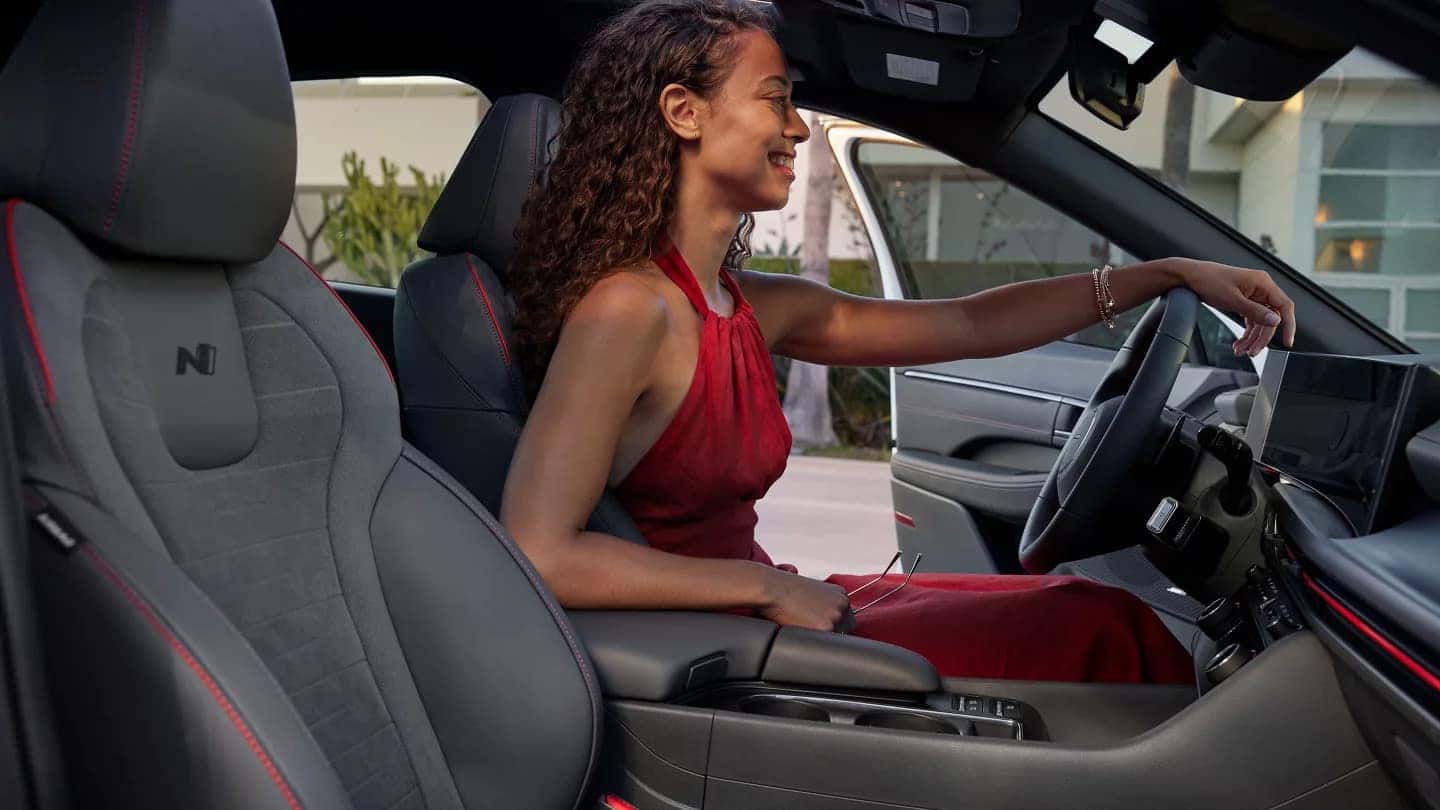 Woman in a red dress sitting in the driver’s seat of the 2024 Hyundai Sonata N Line, smiling and holding sunglasses.