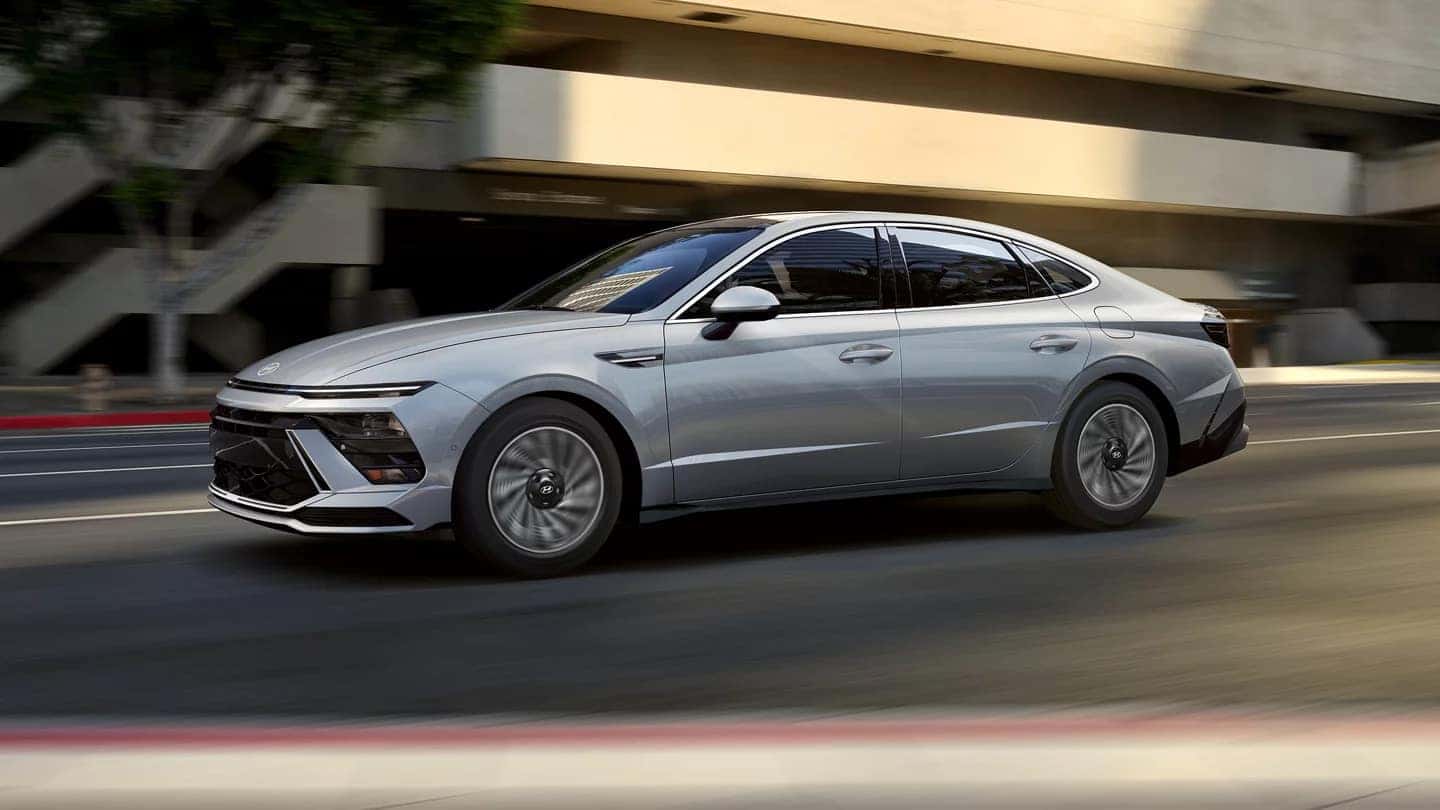 A silver 2024 Hyundai Sonata Hybrid driving on a city street, showcasing its streamlined design and futuristic lighting signature.