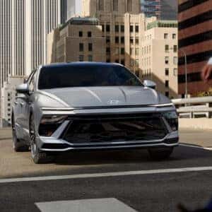 A bold front-angle shot of the silver 2024 Hyundai Sonata Hybrid driving through a city with pedestrians in the foreground.